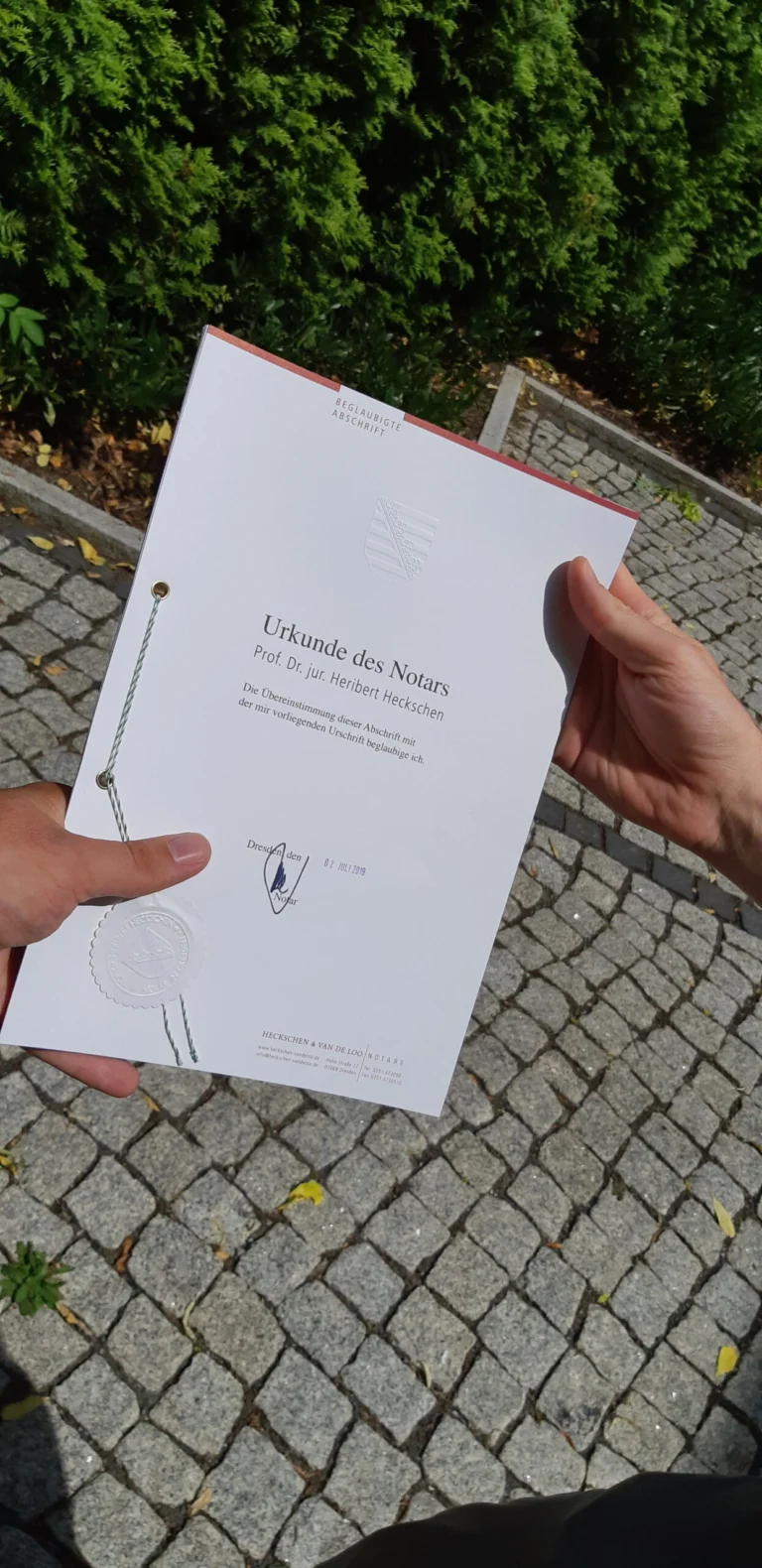 Gründungsurkunde wird im Sonnenlicht vor einer grünen Hecke gehalten.