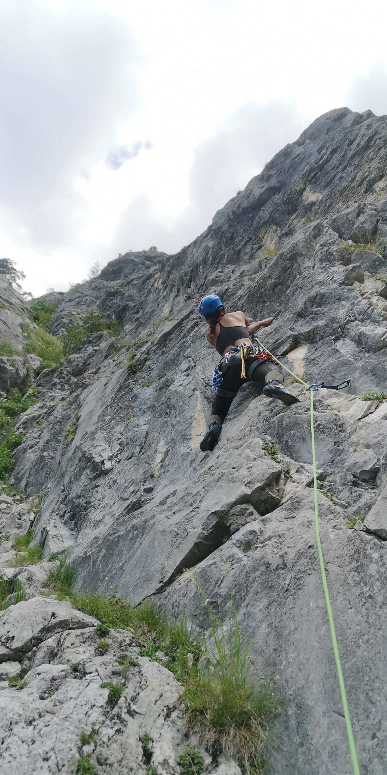 Person beim Felsklettern mit Kletterausrüstung an steiler Felswand unter wolkigem Himmel.
