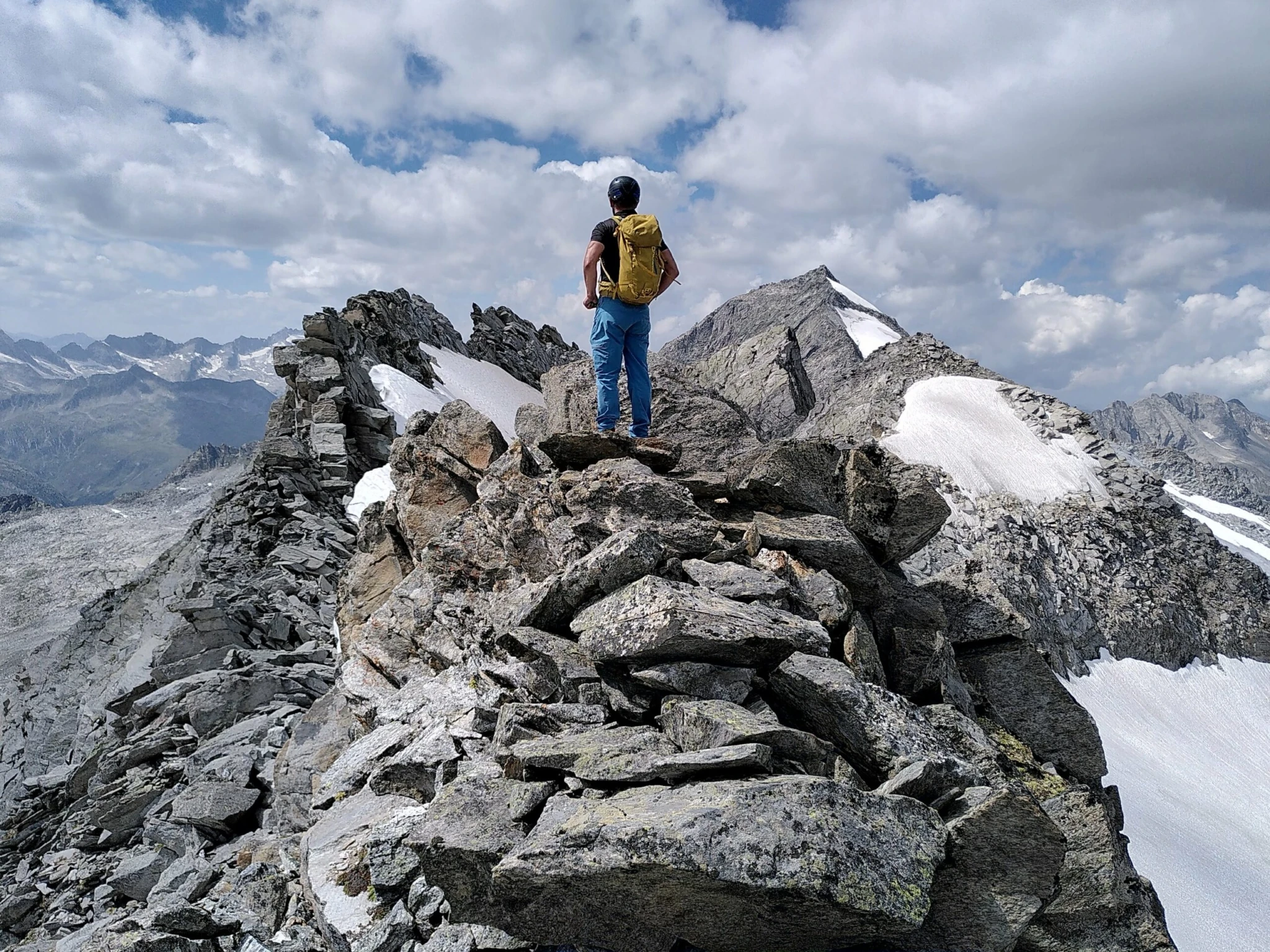 Person steht mit Rucksack auf einem schmalen Felsgrat mit Blick auf schneebedeckte Berge und dramatischen Wolkenhimmel.